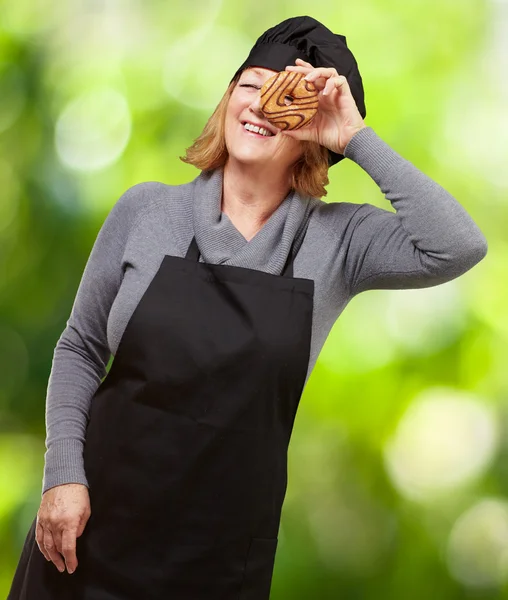 Middle aged cook woman looking through a donut against a nature — Stock Photo, Image