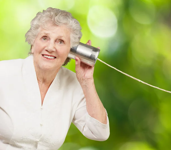 Porträt einer Seniorin, die mit Blechbüchse gegen einen Baum hört — Stockfoto