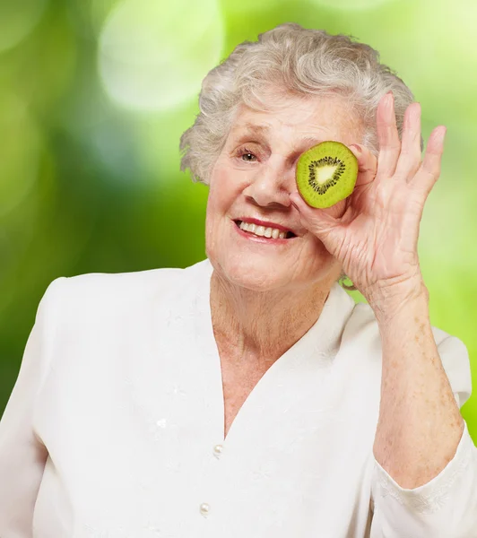 Portrait of senior woman holding kiwi in front of her eye agains — Stock Photo, Image