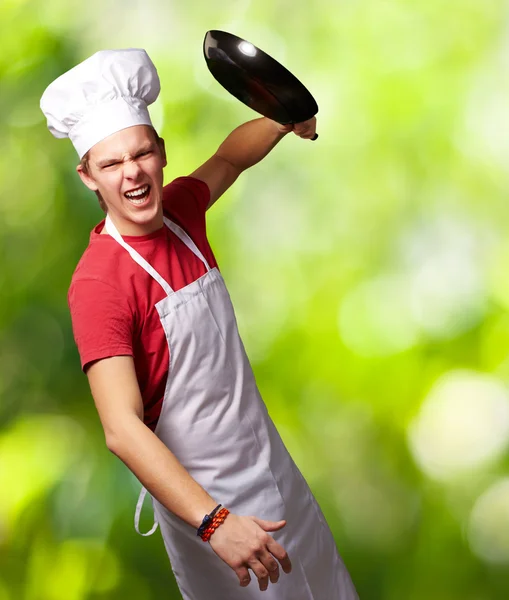 Retrato del joven cocinero enojado golpeando con la sartén contra un natu — Foto de Stock