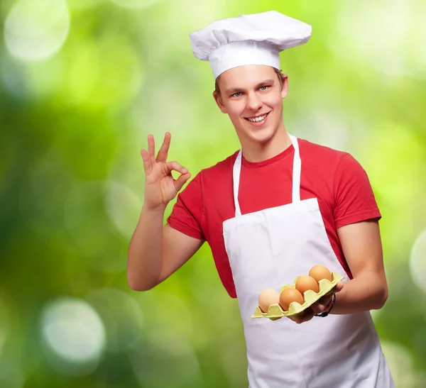Portrait of young cook man holding egg box and doing good gestur — Stock Photo, Image