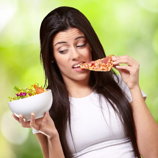 Retrato de una mujer joven comiendo pizza y mirando ensalada contra un —  Fotos de Stock