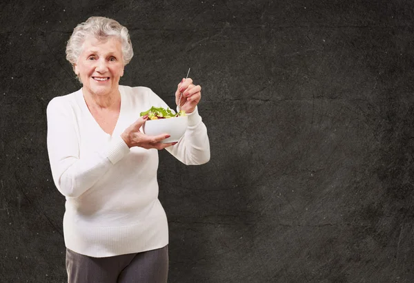 Portrait of senior woman eating salad against a grunge wall — Stock Photo, Image