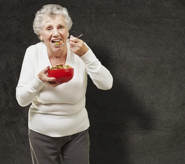 Senior woman eating cereals out of a red bowl against a grunge b — Stock Photo, Image
