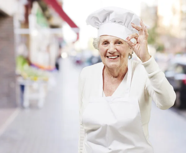 Старшая женщина повар делает отличный символ против улицы bac — стоковое фото