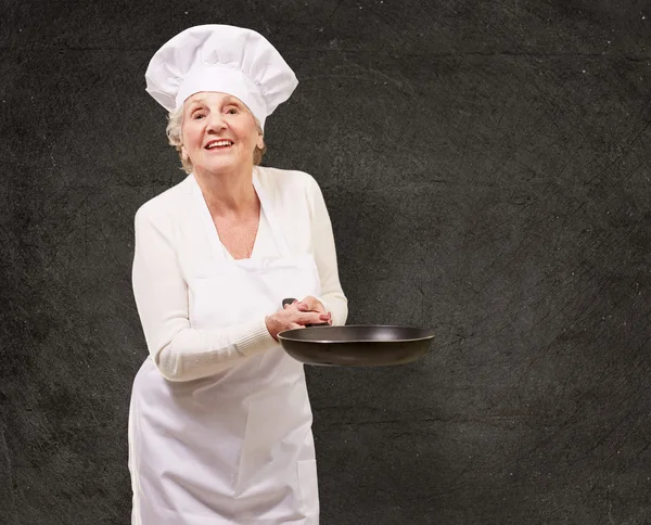 Portrait of a friendly cook senior woman holding pan against a g — Stock Photo, Image