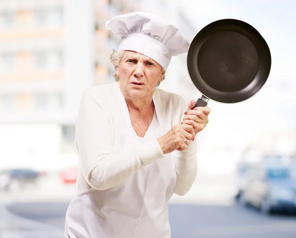 stock image Cook senior woman angry trying to hit with pan against a buildin