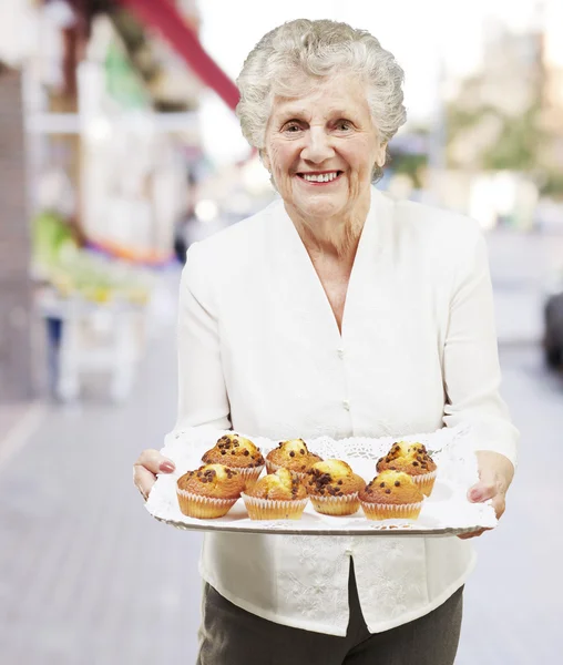 Mujer mayor sonriendo y sosteniendo una bandeja con magdalenas en la calle — Foto de Stock