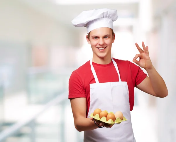 Retrato de un joven cocinero sosteniendo una caja de huevos y haciendo un buen gesto — Foto de Stock