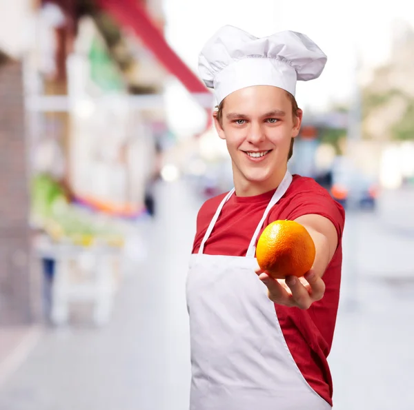 Portrait of young cook man offering orange at street — Stock Photo, Image