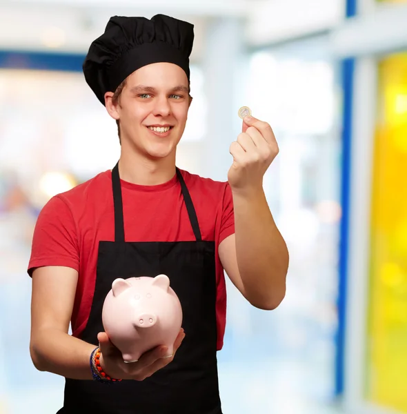 Retrato de jovem cozinheiro segurando moeda de euro e banco porquinho indo — Fotografia de Stock