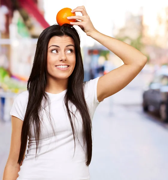 Portrait of young woman holding orange on her head at city — Stock Photo, Image