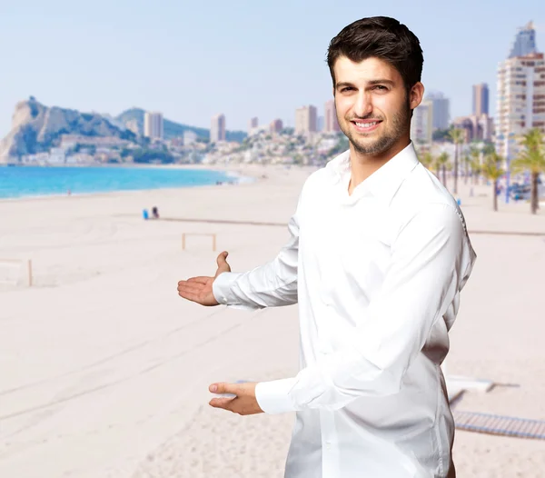 Portrait of young man doing welcome gesture against a beach — Stock Photo, Image