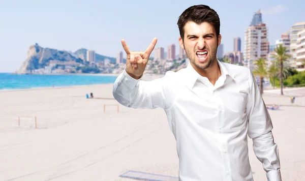 Porträt eines jungen Mannes, der Rock-Symbol gegen einen Strand macht — Stockfoto