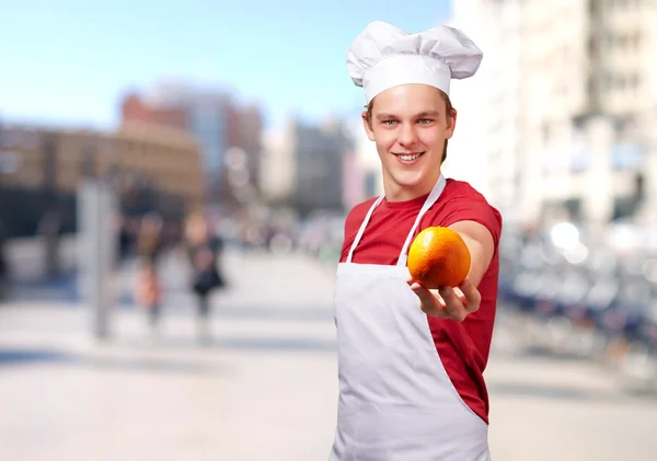 Portrait de jeune cuisinier homme offrant orange à la ville — Photo