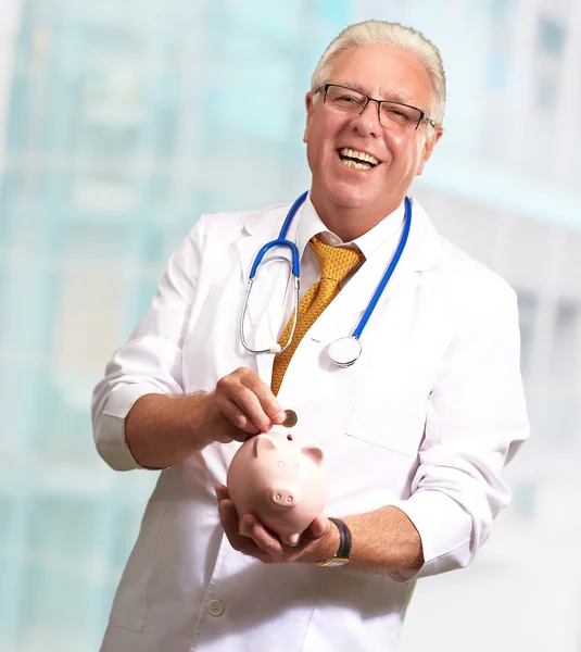 Médico masculino colocando moedas em um banco porquinho — Fotografia de Stock