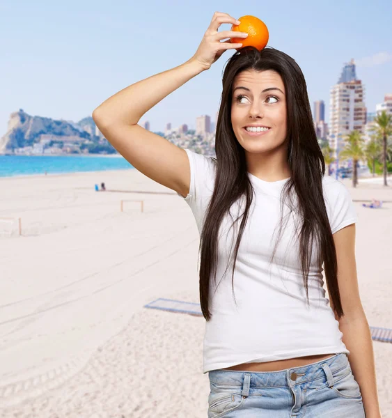 Portrait of young woman holding orange on head against a beach — Stock Photo, Image