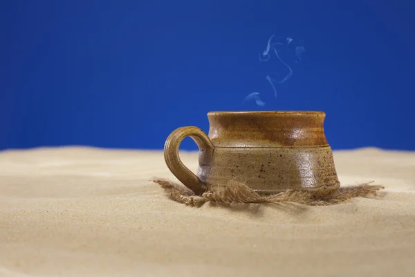 Clay mug with tea or coffee on beach sand, blue background