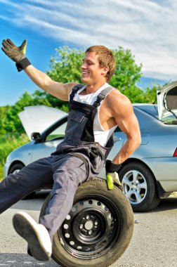 Portrait of laughing mechanic sitting on a tire on a road. clipart