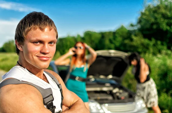 Portrait of handsome mechanic with two helpless women and broken car on background — Stock Photo, Image