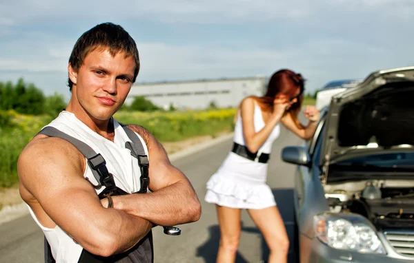 Porträtt av en hadsome mekaniker med en tjej nära trasiga bil på en bakgrund — Stockfoto