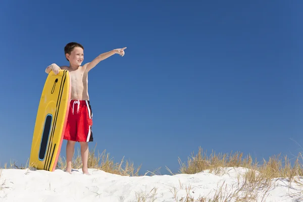Ung pojke barn på en strand med surfbräda pekar — Stockfoto