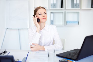 Beautiful young business woman sitting at office desk and talkin clipart