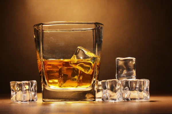 Glass of scotch whiskey and ice on wooden table on brown background — Stock Photo, Image
