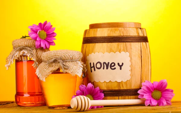 Süßer Honig in Fässern und Gläsern mit Nieselregen auf Holztisch auf gelbem Hintergrund — Stockfoto