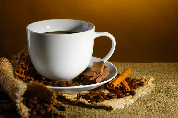 Xícara de café e feijão, paus de canela e chocolate no saque no fundo marrom — Fotografia de Stock