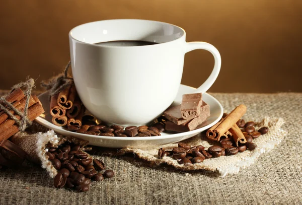 Cup of coffee and beans, cinnamon sticks and chocolate on sacking on brown background — Stock Photo, Image