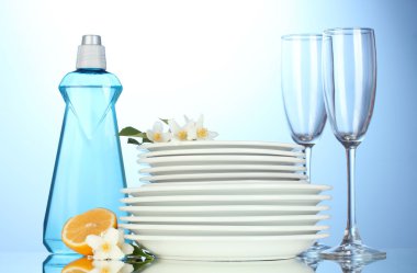 Empty clean plates and glasses with dishwashing liquid, sponges and lemon on blue background clipart