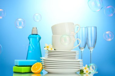 Empty clean plates, glasses and cups with dishwashing liquid, sponges and lemon on blue background clipart