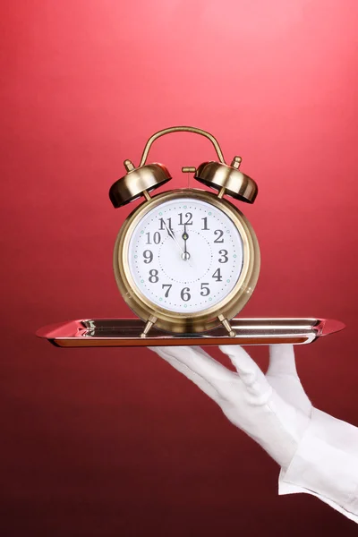 लाल पृष्ठभूमि पर अलार्म घड़ी के साथ चांदी की ट्रे पकड़ने वाले दस्ताने में हाथ — स्टॉक फ़ोटो, इमेज