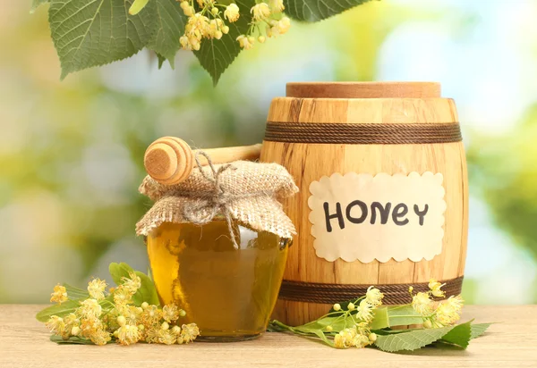 Jar and barrel with linden honey and flowers on wooden table on green background — Stock Photo, Image