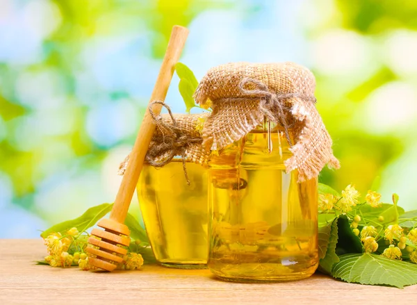 Jars with linden honey and flowers on wooden table on green background — Stock Photo, Image