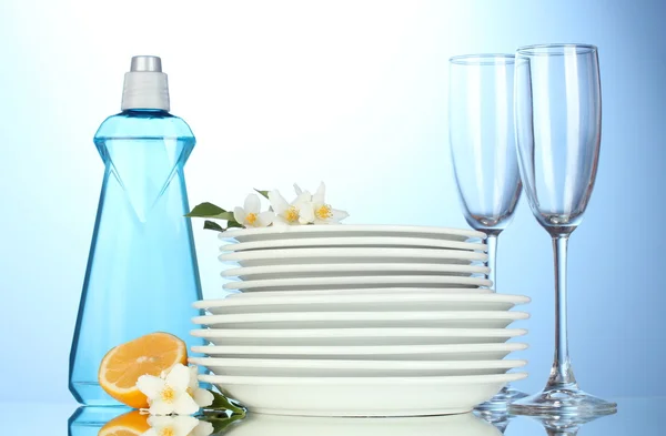 Piatti puliti vuoti e bicchieri con detersivo per piatti, spugne e limone su fondo blu — Foto Stock