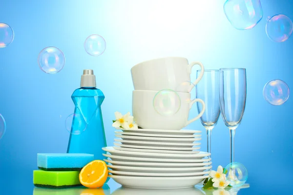 Пустые чистые тарелки, стаканы и чашки с жидкостью для мытья посуды, губки и лимон на синем фоне — стоковое фото