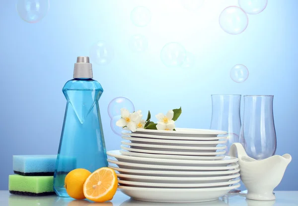 Platos y vasos limpios vacíos con líquido lavavajillas, esponjas y limón sobre fondo azul —  Fotos de Stock
