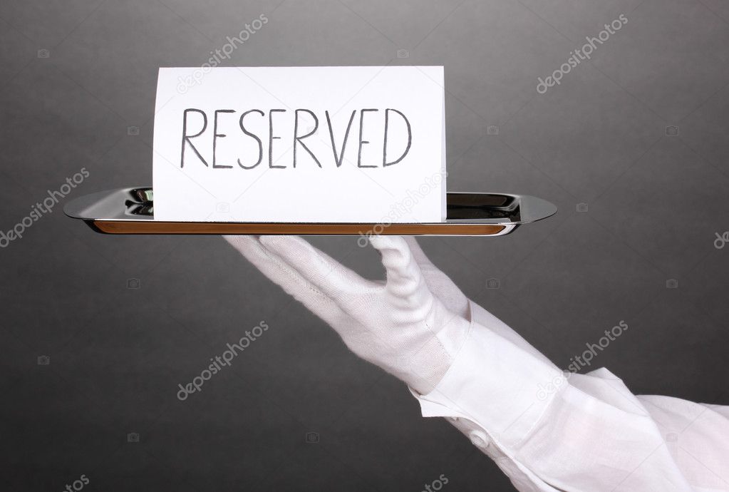 Hand in glove holding silver tray with card saying reserved on grey background