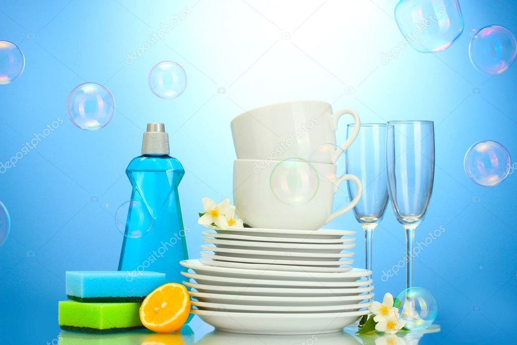 Empty clean plates, glasses and cups with dishwashing liquid, sponges and lemon on blue background