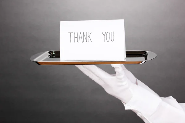 Mão na luva segurando bandeja de prata com cartão dizendo obrigado no fundo cinza — Fotografia de Stock