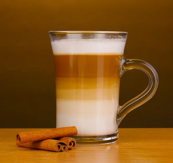 Fragrant сappuccino latte in glass cup and cinnamon on wooden table on brown background — Stok Foto