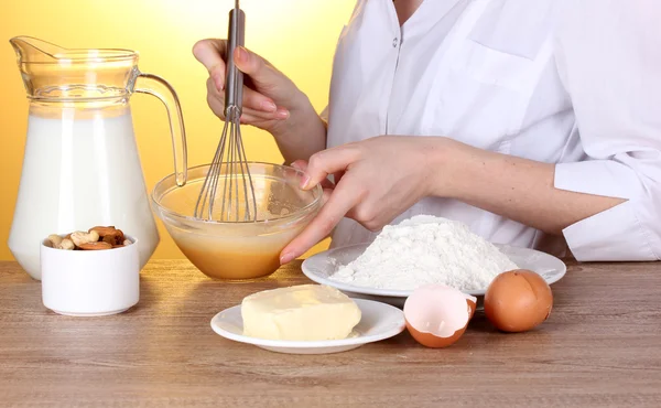 Женские руки смешивают яйца в миске на деревянном столе на желтом фоне — стоковое фото