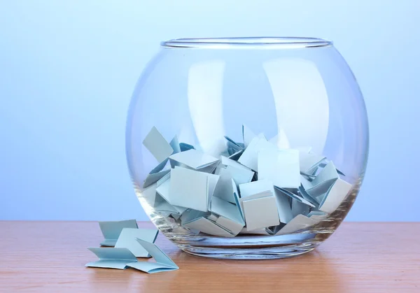 Kousky papíru pro loterie v váza na dřevěný stůl na modrém pozadí — Stock fotografie
