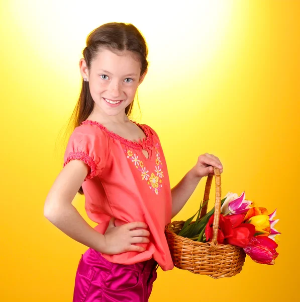 Portrait of beautiful little girl with tulips in basket on yellow background — Stock Photo, Image