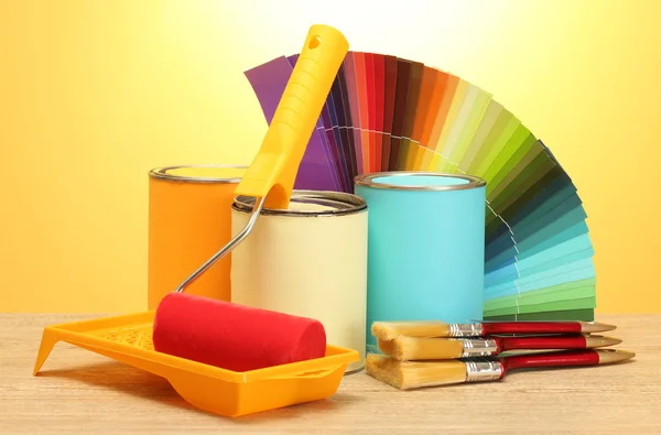 Tin cans with paint, roller, brushes and bright palette of colors on wooden table on yellow background — Stock Photo, Image