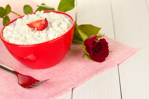 Requesón con fresa en tazón rojo, tenedor y flor en servilleta rosa sobre mesa de madera blanca de cerca —  Fotos de Stock
