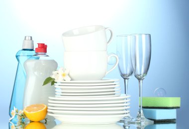 Empty clean plates, glasses and cups with dishwashing liquid, sponges and lemon on blue background clipart