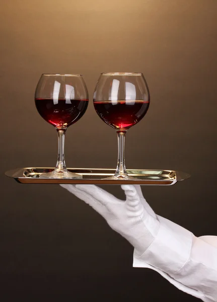 Hand in glove holding silver tray with wineglasses on brown background